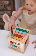 A toddler in a light pink dress plays with the wooden activity cube by Mentari, using a red mallet to tap the colorful xylophone on top.