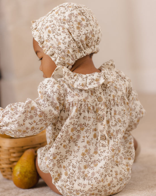 A baby wearing a cream-colored floral romper and matching bonnet, sitting on a carpeted floor next to a wicker basket and a pear.