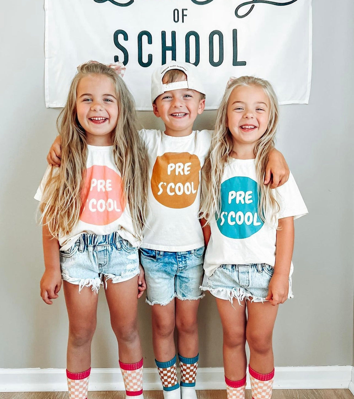 Three children standing together indoors wearing matching white graphic T-shirts with colorful circles and the words 'PRE S COOL,' paired with denim shorts and patterned socks, in front of a wall banner that reads 'LOVE OF SCHOOL.'.