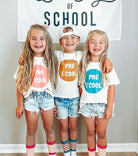 Three children standing together indoors wearing matching white graphic T-shirts with colorful circles and the words 'PRE S COOL,' paired with denim shorts and patterned socks, in front of a wall banner that reads 'LOVE OF SCHOOL.'.