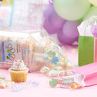 A styled tabletop scene with pastel party decorations, including balloons, a pink gift bag, a cupcake, and several clear jars and bags filled with colorful candy and wrapped sweets on a pink surface.