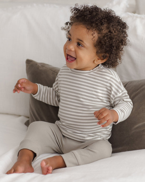 A baby with curly dark hair sits smiling on a white bed, wearing a gray and white striped long-sleeve top and light beige pants.