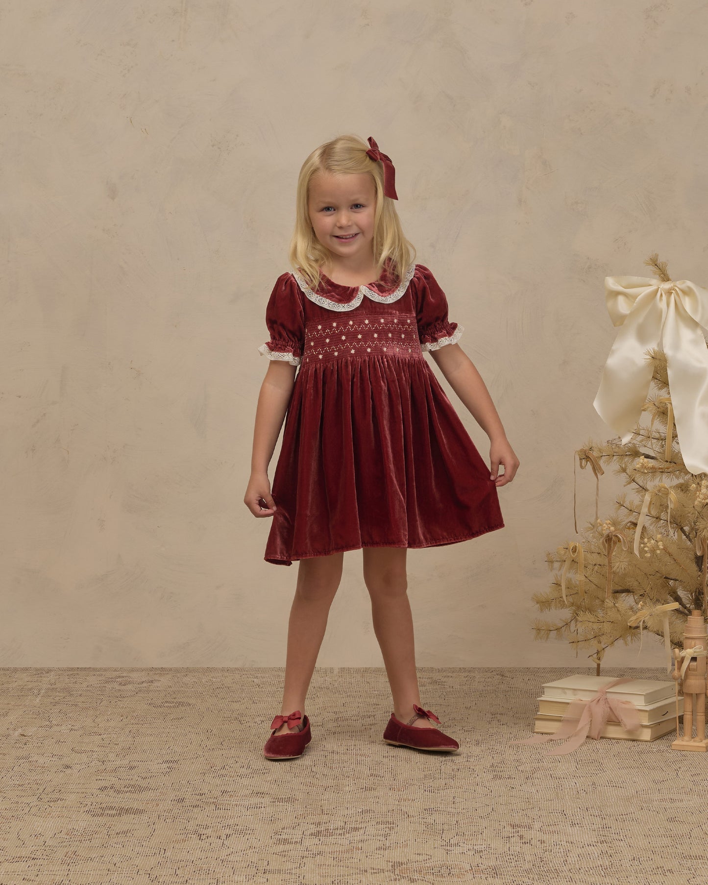A young girl stands in a studio wearing a burgundy velvet short-sleeve dress with white embroidered trim around the collar and cuffs, paired with matching burgundy shoes. The background is a textured beige wall and floor, with a decorative dried tree and books on the right.
