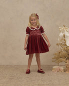 A young girl stands in a studio wearing a burgundy velvet short-sleeve dress with white embroidered trim around the collar and cuffs, paired with matching burgundy shoes. The background is a textured beige wall and floor, with a decorative dried tree and books on the right.