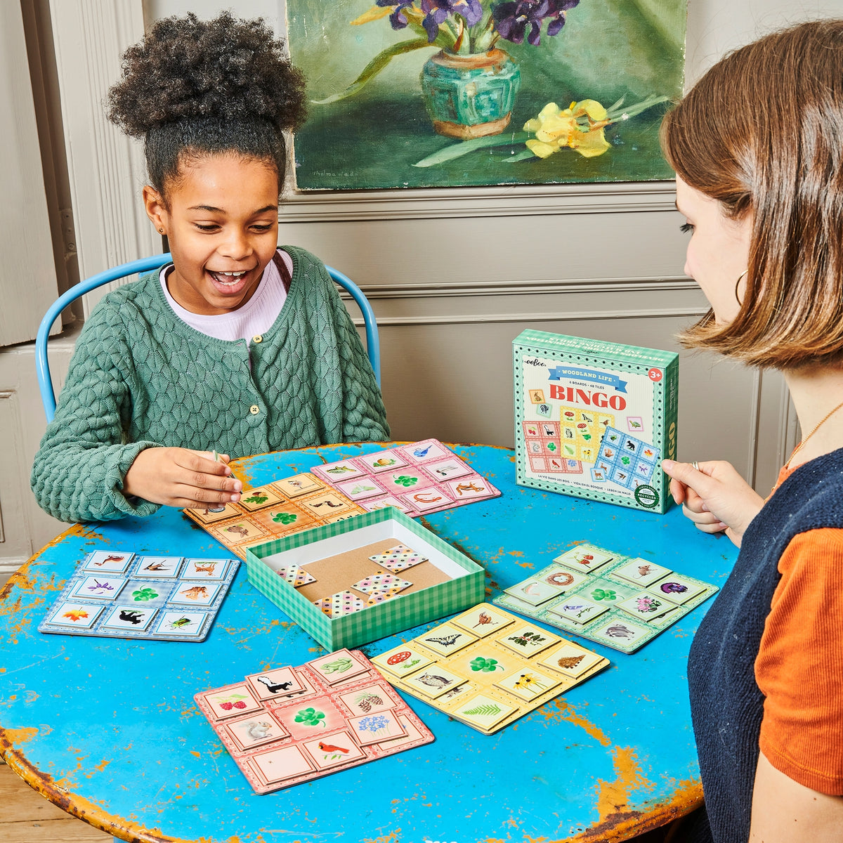Two children playing a board game at a table with colorful cards and a Bingo box.