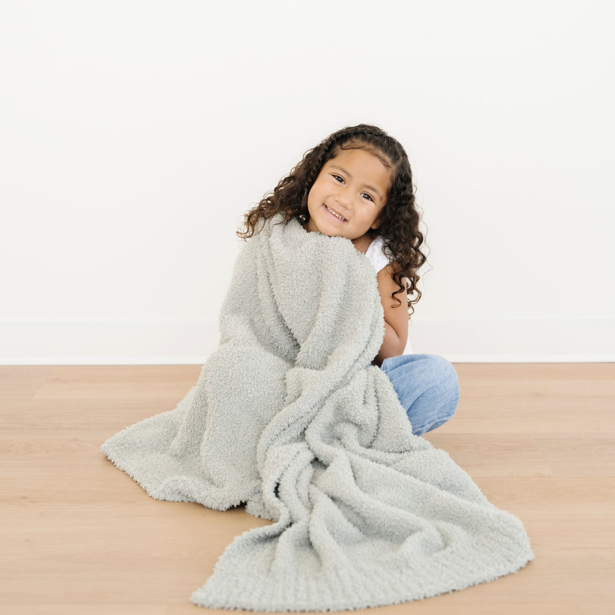 Child wrapped in a cozy gray blanket on a wooden floor with a white background