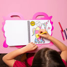 Child coloring a unicorn on a pink table with coloring book and markers.