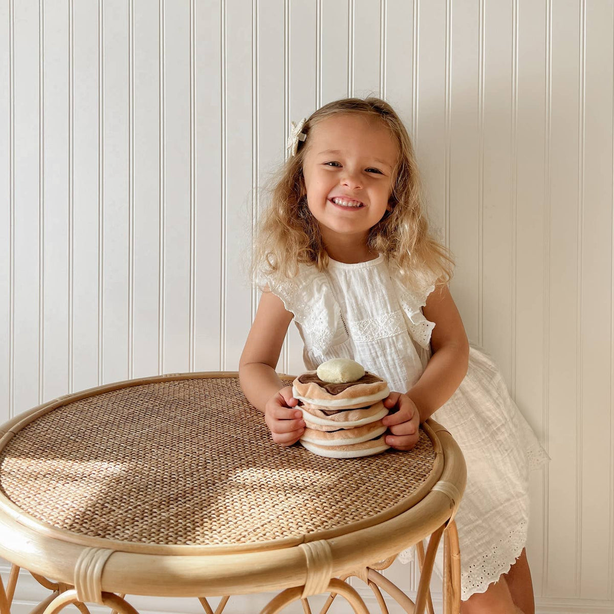 Young girl in a white dress holding a stack of plates on a small round table against a light-colored wall.