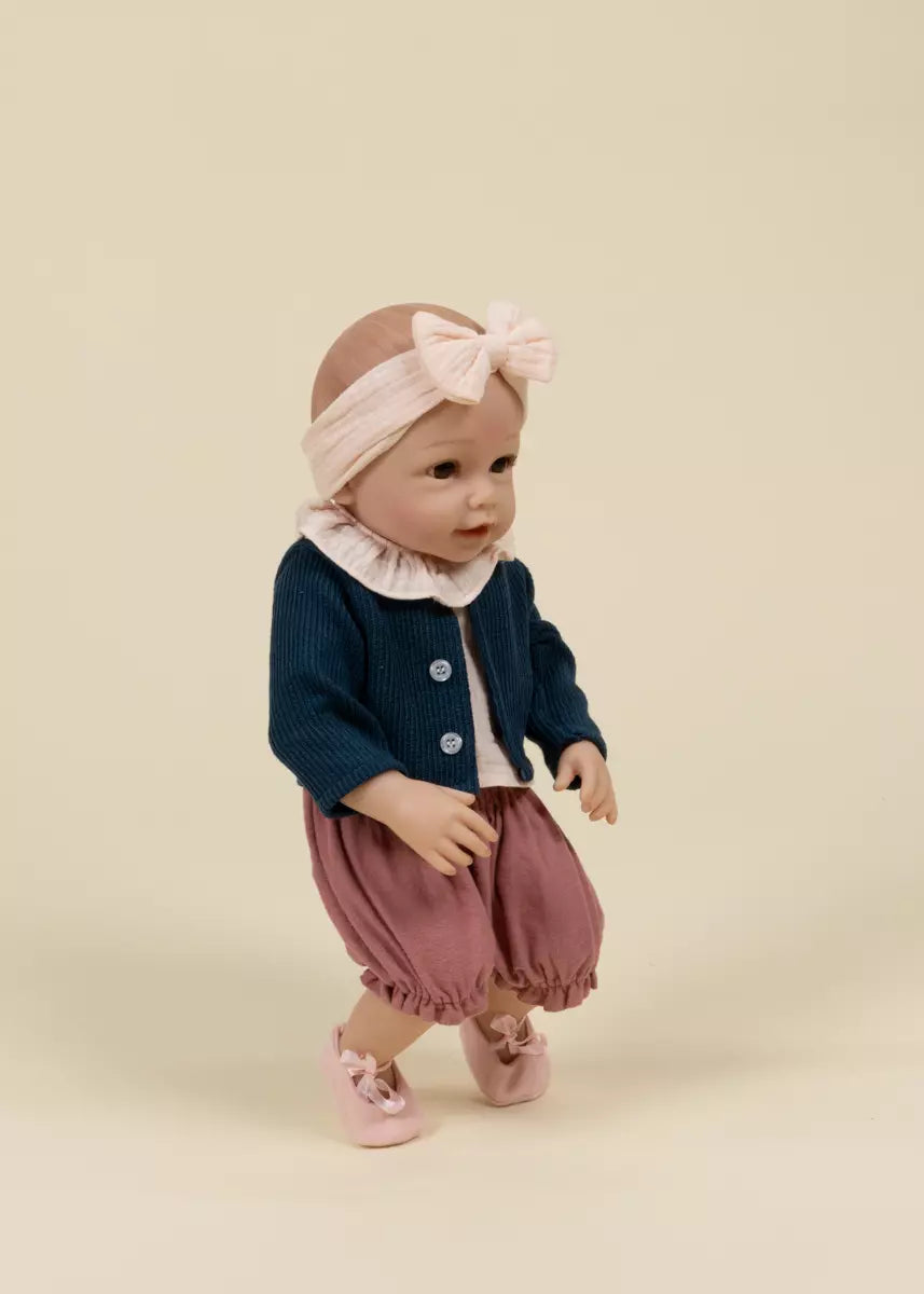 Baby doll wearing a navy cardigan, pink pants, and a headband on a beige background