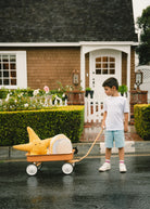 Child pulling a wagon with a large toy dinosaur in front of a house.