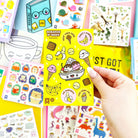 Hand holding a yellow scratch and sniff sticker book with various stickers on a colorful background