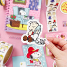 Hand holding a Peanuts character sticker with a colorful background of more stickers and a toy house.