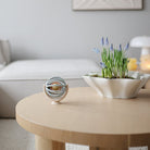 A blue and beige Mushie Suction Spinner Ball sitting on a round wooden coffee table in a living room, with a white bowl of plants and a sofa in the background.