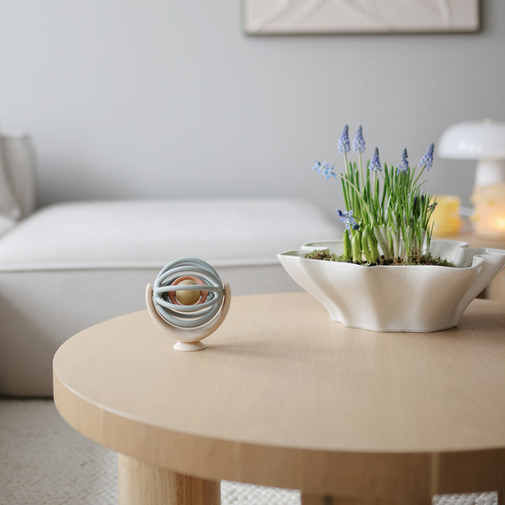 A blue and beige Mushie Suction Spinner Ball sitting on a round wooden coffee table in a living room, with a white bowl of plants and a sofa in the background.