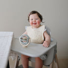 A smiling baby sitting in a wooden high chair, looking at and touching a blue and beige Mushie Suction Spinner Ball attached to the tray.