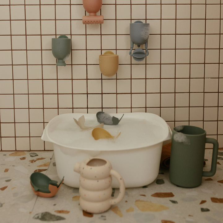 A speckled ceramic teapot and a green pitcher resting on a terrazzo surface near a large white basin filled with water and floating toy whales, set against a green-tiled wall with wall-mounted ceramic decor.