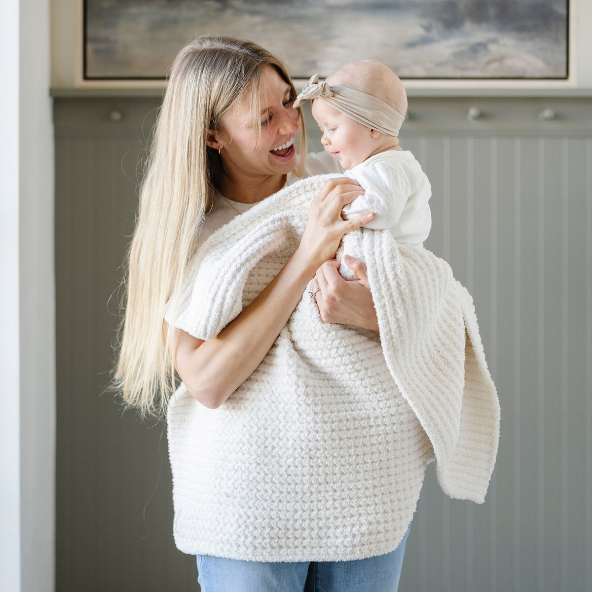 Woman holding a baby wrapped in a white blanket indoors.