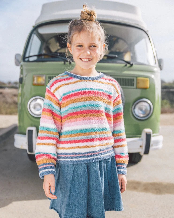 A young girl smiling in front of a vintage green van, wearing a fuzzy long-sleeve sweater with colorful horizontal stripes in pink, orange, yellow, green, blue, and purple, paired with a blue denim skirt.