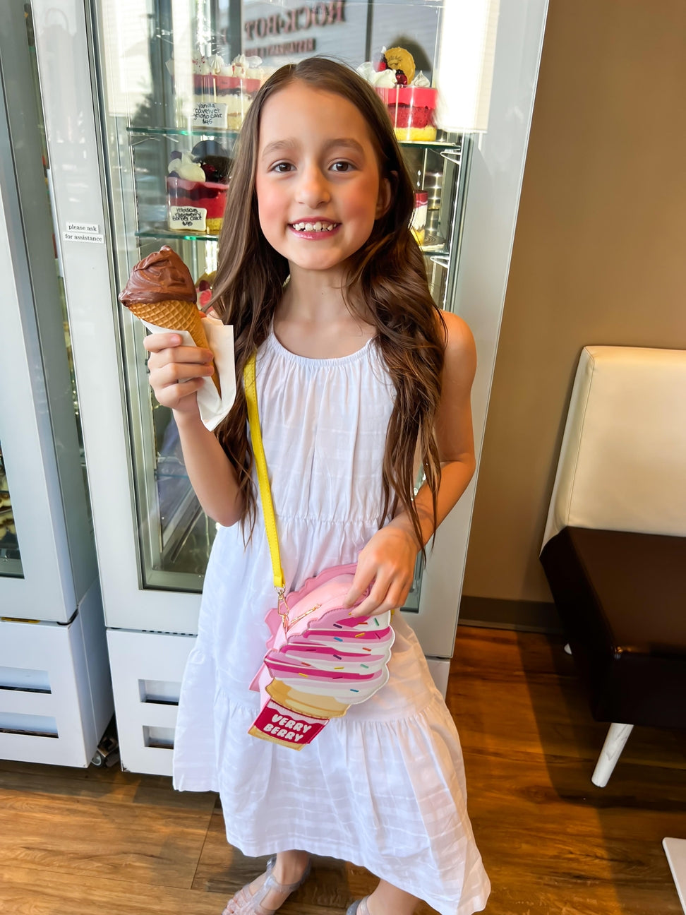 Young girl in a white dress holding a chocolate ice cream cone and a pink bag with a donut design.