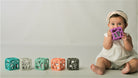 A baby wearing a white dress and headband sits on the right holding a purple cube toy, with five colorful geometric baby toys lined up on the left against a plain light gray background.