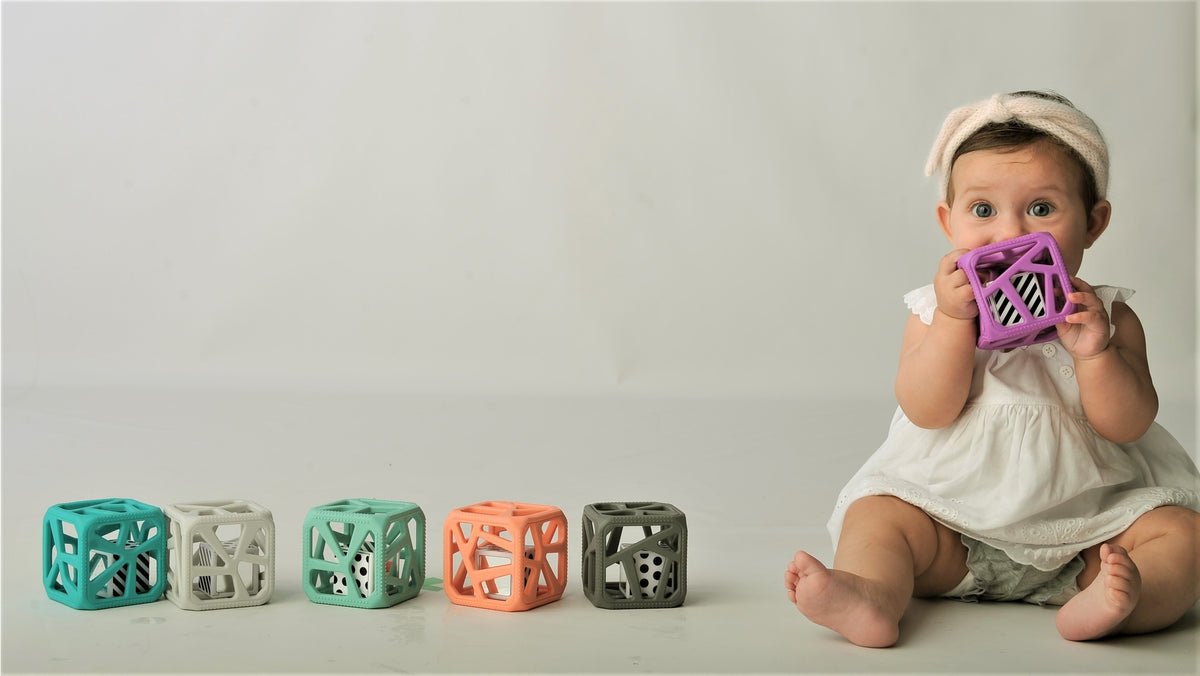 A baby wearing a white dress and headband sits on the right holding a purple cube toy, with five colorful geometric baby toys lined up on the left against a plain light gray background.