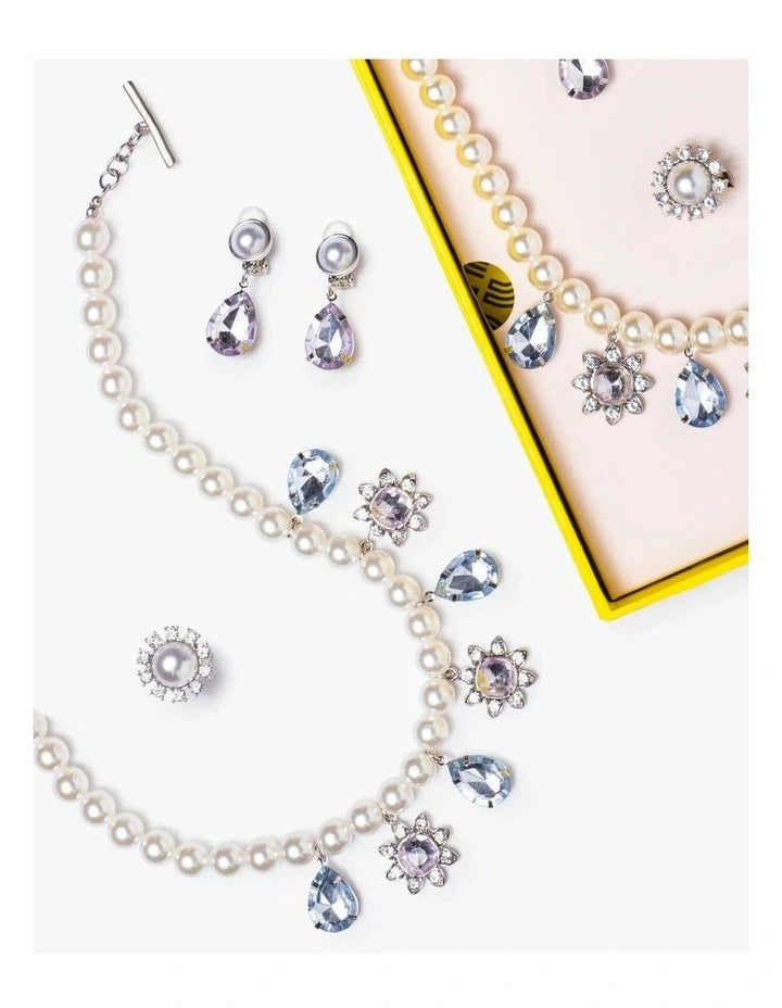 A pearl jewelry set arranged on a white background, including a necklace with teardrop and flower-shaped blue crystal accents, matching earrings, and a partial matching necklace displayed inside a pink box with a yellow border.