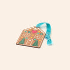A gingerbread house shaped ornament with a turquoise ribbon loop, decorated with colorful icing-like patterns and small tree motifs, shown on a plain light beige background.