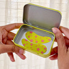 Hands holding a small open tin with yellow and pink star-shaped objects inside.