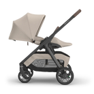A stroller in a light beige and dark gray/black colorway, shown from a side profile on a clean white background. The stroller features a beige canopy, a matching seat liner, a dark gray frame, and brown accents on the handle grip.