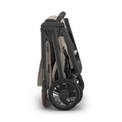A folded stroller shown from the side on a clean white background. The stroller features a dark gray frame, beige fabric body, and brown accents on the handle and wheel.