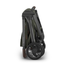A folded stroller shown from the side on a clean white background. The stroller features an olive green fabric body with a dark gray frame and black wheels.