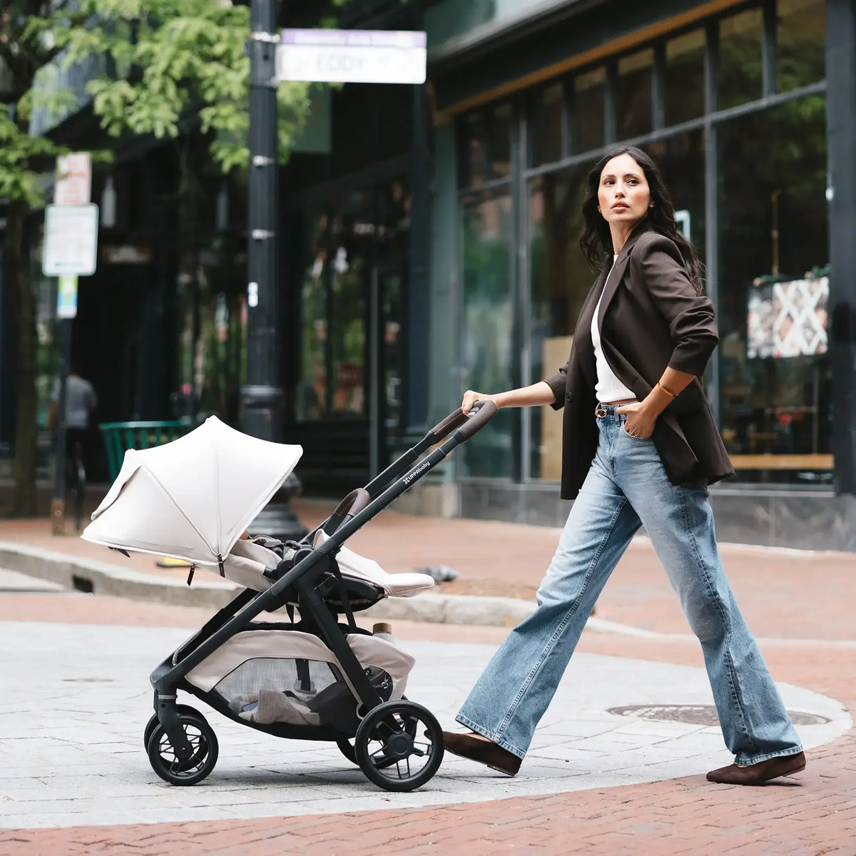 A woman walks on a city sidewalk pushing a stroller. She is wearing a dark brown blazer, white top, and light blue flared jeans.