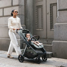 A woman in a cream knit sweater and white pants pushes a dark green stroller with a baby inside, walking along a stone sidewalk in front of a gray building.