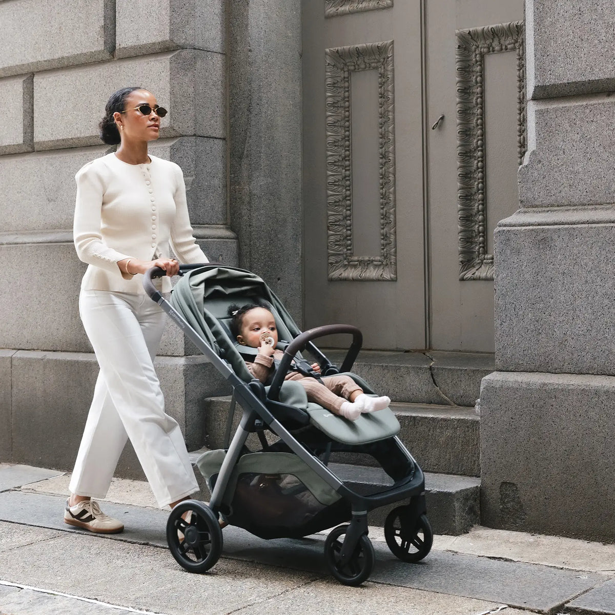 A woman in a cream knit sweater and white pants pushes a dark green stroller with a baby inside, walking along a stone sidewalk in front of a gray building.