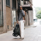 A woman carrying a child walks on a city sidewalk while pulling a black stroller. She is wearing a dark gray long-sleeve top and white wide-leg pants.