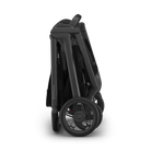 A folded stroller shown from the side on a clean white background. The stroller features an olive green fabric body with a dark gray frame and black wheels.