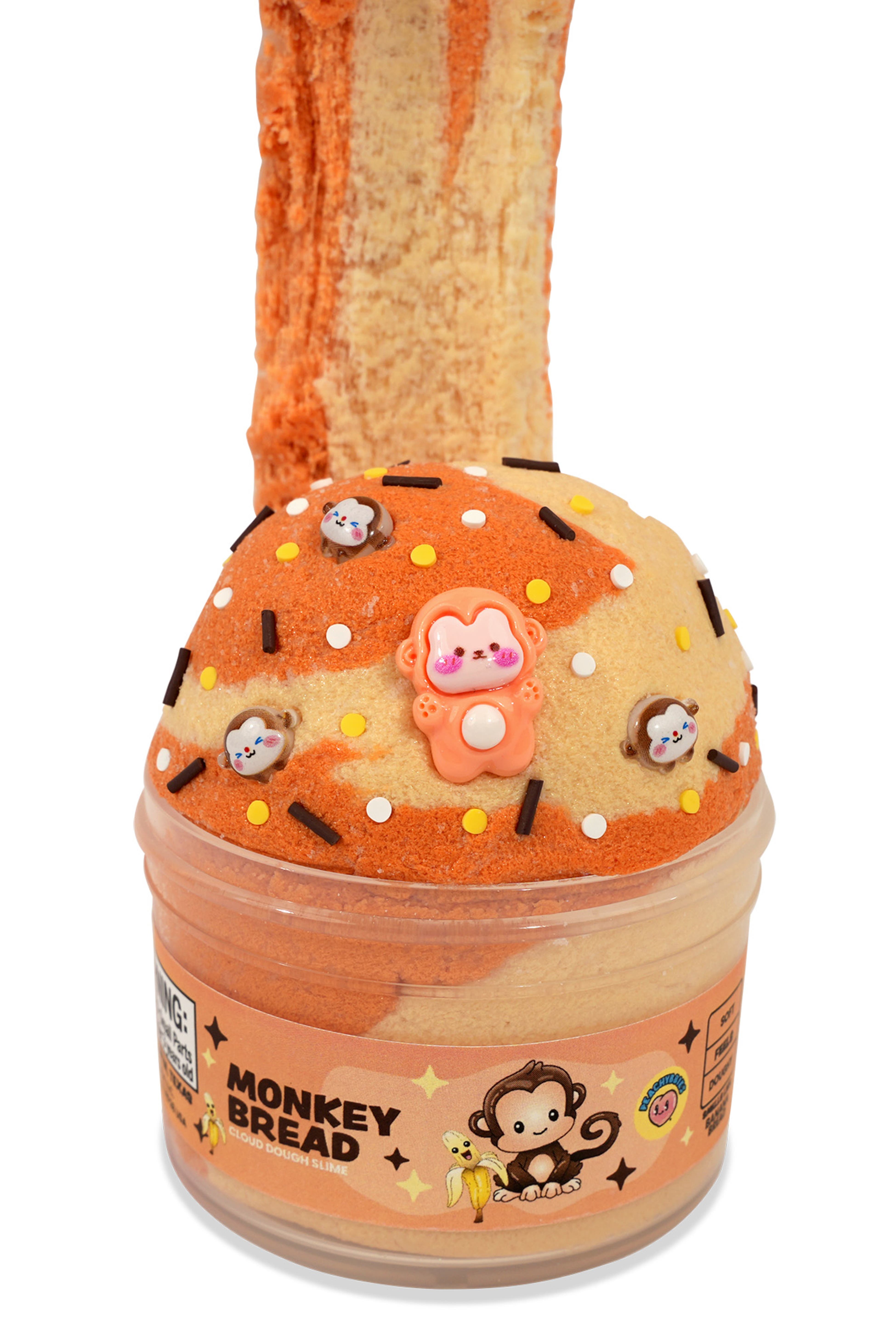A novelty slime jar shaped like a donut, labeled 'MONKEY BREAD' on the front. The jar contains orange and beige marbled slime, topped with a beige donut-slice shape decorated with small monkey charms, white and black sprinkles, and a central pink monkey figure.