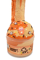 A novelty slime jar shaped like a donut, labeled 'MONKEY BREAD' on the front. The jar contains orange and beige marbled slime, topped with a beige donut-slice shape decorated with small monkey charms, white and black sprinkles, and a central pink monkey figure.