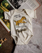 An off-white short-sleeve baby bodysuit laid flat on a textured surface, featuring a graphic of a yellow and white puppy with the text 'BRING ME ALL THE Puppies'. The bodysuit is styled with vintage books and a small wooden toy.