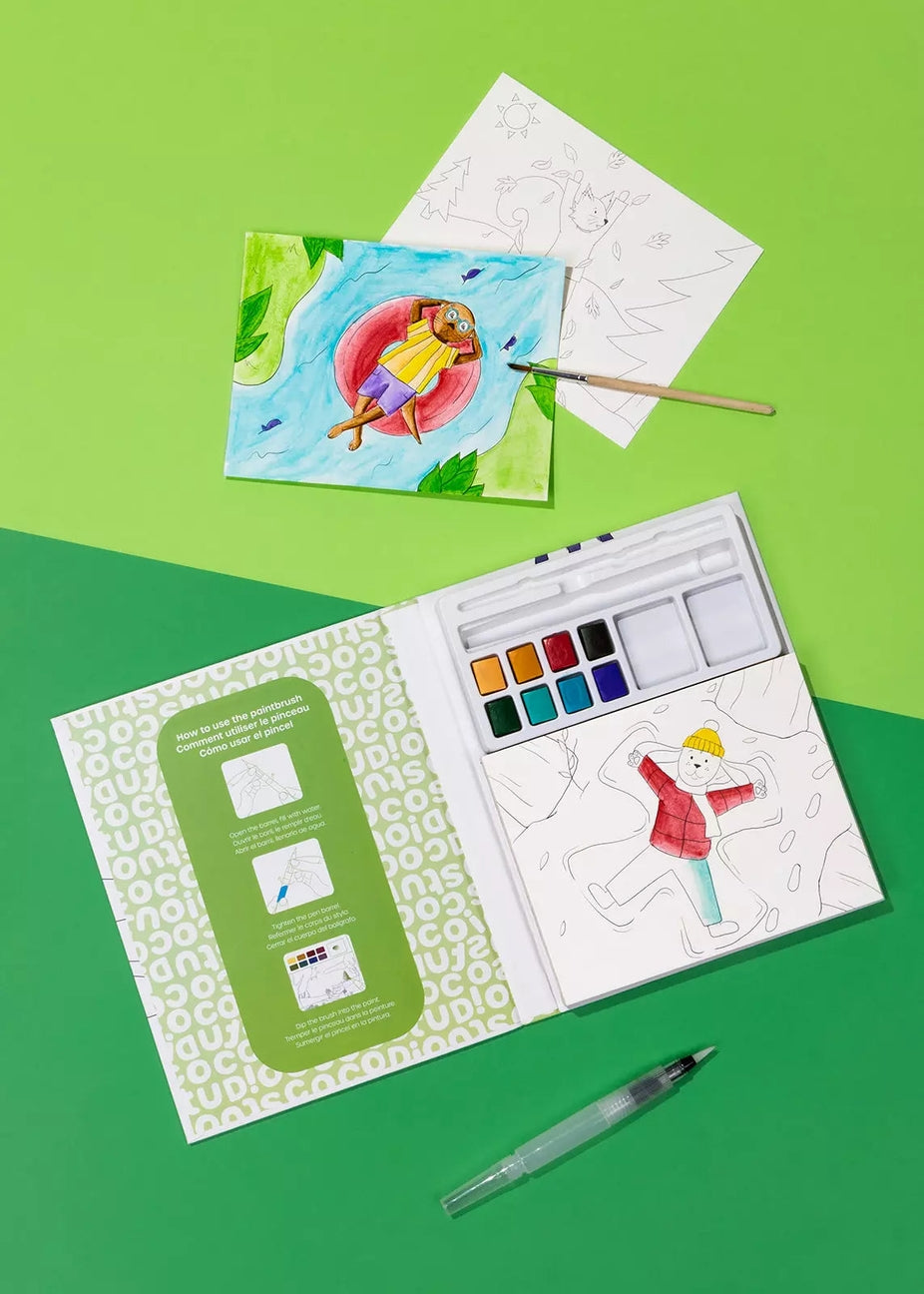 An open children's watercolor painting kit on a bright green background, with a painted sample card, a paintbrush, a pencil, and a water pen arranged around it. The kit includes a palette, paper, and printed instruction panel.