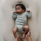 Baby in a light green onesie lying on a white cushion, held by an adult's hands.