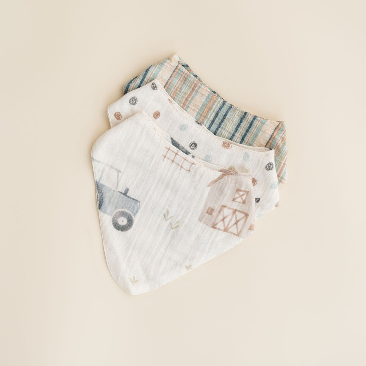 Two reusable muslin baby bibs stacked on a warm beige background. The front bib is white with a rustic farm-themed print including a tractor, barn, and fence in muted gray and brown.