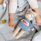 Children's legs with a teddy bear wearing a 'SKI' sweater on a textured blanket.