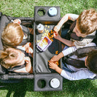 Children sitting in strollers around a portable dining table with food on a grassy surface
