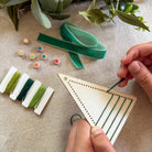 Person threading green yarn through a wooden loom with embroidery floss and wooden beads on a textured surface.