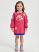A young girl stands against a light gray studio background wearing a bright pink fuzzy long-sleeve dress with a rainbow and flower applique design, blue and pink trim, and pink boots.