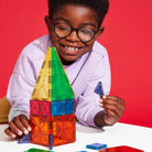 A smiling child wearing glasses and a light purple sweatshirt builds with colorful translucent magnetic tiles, holding a small blue piece in one hand while a tall tower structure stands on the table in front of them against a vibrant red background.