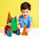 A young child smiling as they play with colorful standard-sized and micro-sized magnetic building tiles on a bright yellow surface.