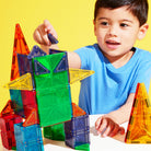 A young child playing on a yellow background, smiling while assembling a colorful tower using standard-sized and micro-sized magnetic building tiles.