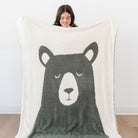 A person holds up a large cream sherpa blanket featuring a dark gray bear face design, with the person's head visible above the blanket against a plain white wall.